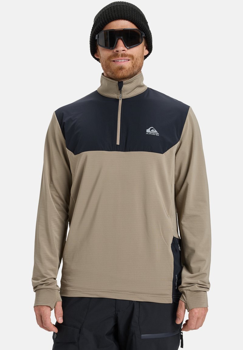 Beige langærmet pullover med en marineblå øverste sektion, halv lynlås, struktureret stof og tommelfingerhuller ved ærmerne, parret med en sort beanie og solbriller.