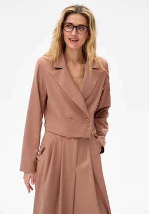 Blondynka w brązowej marynarce z podwójnym rzędem guzików i dopasowanych spodniach z szerokimi nogawkami o wysokim stanie, uśmiechająca się i patrząca w bok.