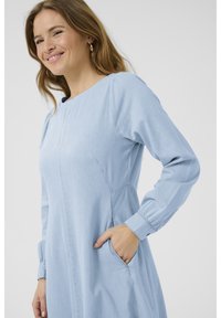 Svetlomodré džínové šaty s dlhými rukávmi, okrúhlym výstrihom, bočnými vreckami a detailmi švov. Mäkka textúra, voľný strih.