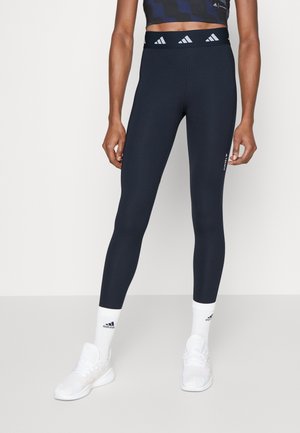 Leggings navy à taille haute, fabriqués en tissu extensible. Présentent un logo discret sur le côté et sont associés à des chaussettes de sport blanches et des chaussures.