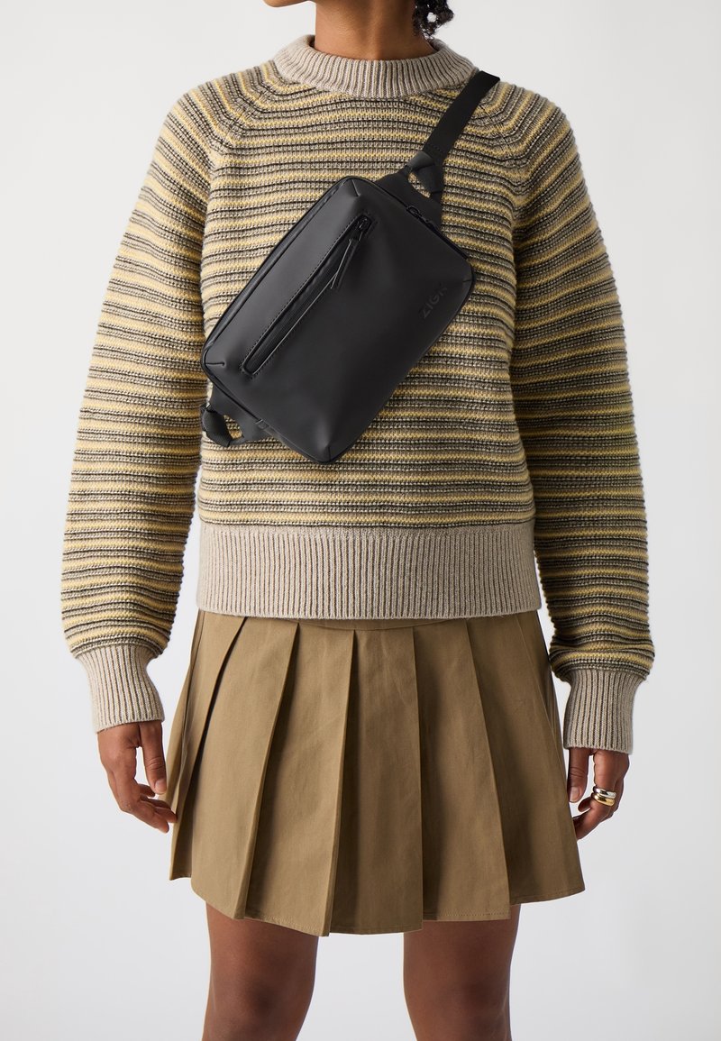 Personne portant un pull en maille rayé et une jupe plissée kaki avec un sac bandoulière noir porté en travers de la poitrine.