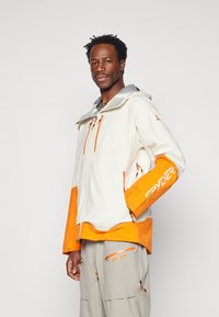 White and orange waterproof jacket with grey hood, zippered pockets, adjustable cuffs, and "SPYDER" logo on the sleeve.