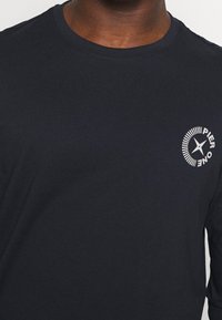 Mörk marinblå crew neck t-shirt med en liten vit cirkulär "Pier One" logo på vänster bröst som bärs av en person.