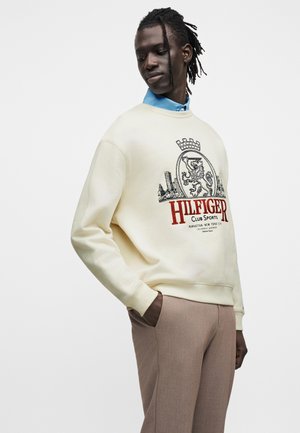 Άνδρας με κρεμ φούτερ με κόκκινο κείμενο "HILFIGER" και έμβλημα λιονταριού, ορατός μπλε γιακάς πουκάμισου, χέρια στις τσέπες μπεζ παντελονιού.