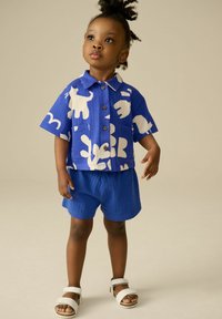 Camicia blu a maniche corte con motivi astratti bianchi, dotata di bottoni e tasca; abbinata a pantaloni corti blu coordinati e sandali leggeri.