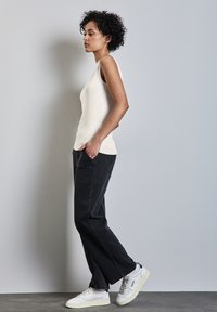 Ivory tank top yang pas dipadukan dengan celana jins hitam longgar, dilengkapi dengan saku depan dan sepatu sneakers putih dengan aksen halus. Latar belakang abu-abu netral.