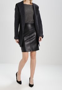 Cardigan en maille noire avec un large col, porté sur un haut rayé gris et une jupe en cuir noir. Stylé avec des chaussures à talons hauts noires.