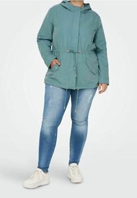 Zelená kapucová bunda s tkaničkou v pase, dvěma předními kapsami, nošená přes bledé modré skinny džíny a bílé tenisky.
