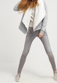 Chaqueta de forro polar gris con un cuello blanco, sobre una blusa de rayas clara, combinada con jeans skinny grises y zapatos tipo slip-on.
