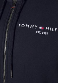 Sudadera con cremallera de color navy, hecha de algodón, con bordado blanco que incluye el nombre de la marca y el año de establecimiento. Cuenta con una cremallera decorativa y cordones.