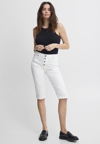 Shorts en denim blanc, style bermuda, avec une taille haute, une fermeture à cinq boutons et des revers, associés à un débardeur noir ajusté et des mocassins noirs.