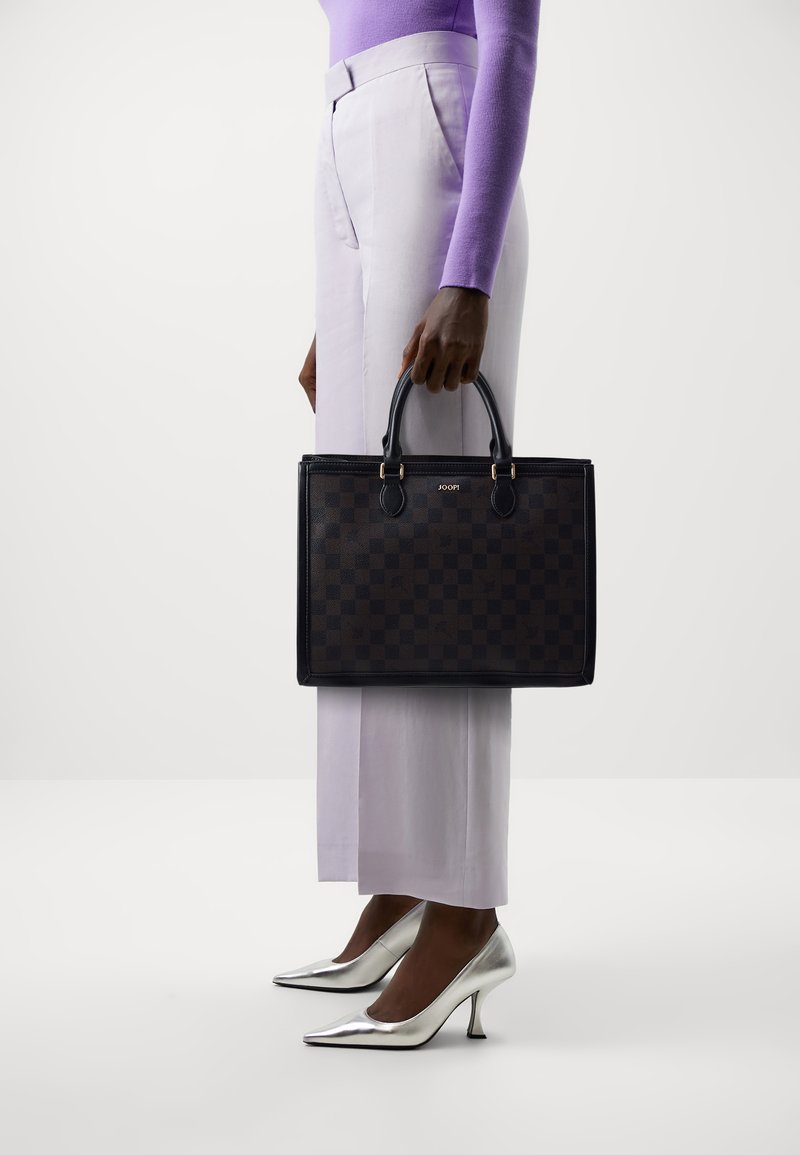 Schwarze Leder-Tote-Tasche mit einem Schachbrettmuster, ausgestattet mit zwei oberen Griffen und goldenen Hardware-Elementen, gehalten neben weiten lavendelfarbenen Hosen.