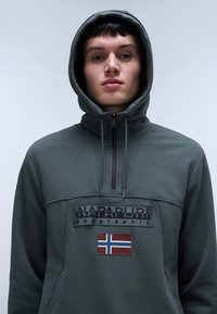 Hoodie verde scuro con zip anteriore, caratterizzato da un patch ricamato e un'emblema della bandiera norvegese. Realizzato in tessuto liscio con una finitura texture.