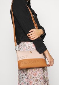 Růžová a béžová crossbody taška s perforovaným detailem "MILUJ SE", hladká kůže, nastavitelný popruh a plochý silueta.