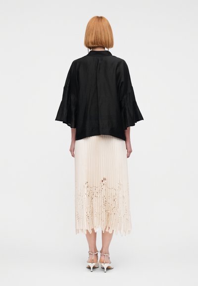 Μαύρο σατέν σακάκι σε στυλ κιμονό με καμπάνες μανίκια πάνω από μια ελαφριά πλισέ φούστα με λουλουδάτες κοψίματα και λευκά πέδιλα με τακούνι.