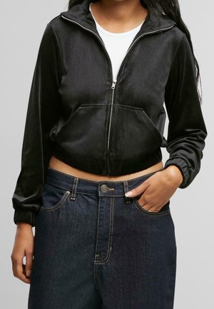 Modèle portant un sweat à capuche court noir zippé par-dessus un haut blanc et un jean foncé, une main dans une poche.