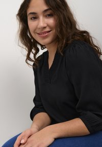 Czarna bluzka z dekoltem w serek, bufkami oraz teksturowymi detalami wzdłuż dekoltu, zestawiona z niebieskimi dżinsami. Miękka tkanina o gładkiej fakturze.