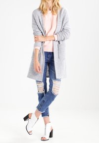 Cárdigan gris de punto con abertura frontal, usado sobre una sudadera rosa. Pantalones vaqueros azules desgastados y botas blancas hasta el tobillo con tacones negros.