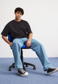 Young man with short dreadlocks sits relaxed on blue office chair, wearing black oversized t-shirt, light blue jeans, and black sneakers.