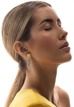 Ženska z zaprtimi očmi, ki nosi rumeno majico in dolge viseče uhane z rumenimi in prozornimi dragimi kamni, lasje sta zvite v nizek konjski rep.