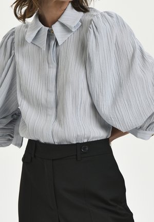 Blouse gris clair texturée avec manches bouffantes et col, associée à un pantalon noir taille haute avec ceinture boutonnée.