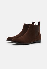 Bottines en daim marron à finition lisse, avec des panneaux élastiques sur les côtés, un bout pointu et un petit talon noir. Tirettes rouges et noires sur le dessus.