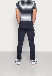 Granatowe spodnie cargo z wieloma bocznymi kieszeniami, zestawione z czarno-białą pasiastą koszulką. Model ma na sobie białe trampki z granatowymi akcentami.