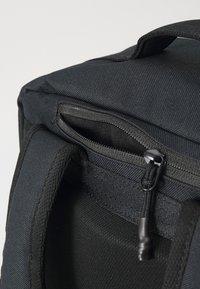 Sac à dos en tissu noir avec des côtés rembourrés, une poignée supérieure et une poche zippée. Caractéristiques d'un matériau texturé et d'une couture durable.