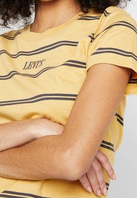 Žluté pruhované tričko vyrobené z bavlny, s námořnickými vodorovnými pruhy a malým logem "Levi's" na přední straně, s viditelnými krátkými rukávy.