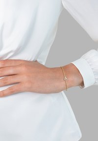 Gold chain bracelet featuring a triangular charm with embedded stones, worn on a wrist against a white blouse background.