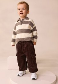 Randig tröja i krämfärgad och grå ull, ihop med bruna kordfluga cargo-byxor. Vita sneakers med grå detaljer, stående på en rund plattform.