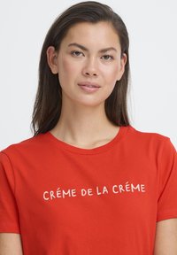 Crvena pamučna majica s kratkim rukavima, okruglim izrezom i glatkom teksturom, s vezanim natpisom "CRÈME DE LA CRÈME" u bijeloj boji.