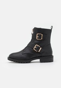 Bottes noires à la cheville en cuir synthétique, avec deux boucles en métal doré et une fermeture éclair sur le côté. Semelle crantée pour une bonne adhérence.