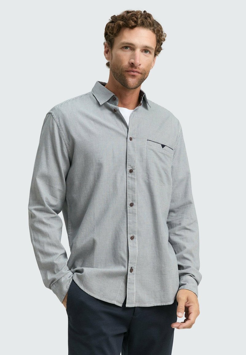 Uomo che indossa una camicia a quadri grigia con taschino e pantaloni scuri, in piedi con una mano in tasca contro uno sfondo semplice.