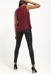 Blusa senza maniche bordeaux con dettaglio cut-out sulla schiena, abbinata a jeans skinny neri e tacchi alti neri, in piedi contro uno sfondo neutro.