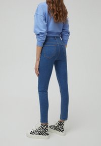 Úzké modré džíny s vysokým pasem, které mají klasický design s pěti kapsami. Modelka má na sobě černobílé pruhované tenisky s vysokou podrážkou.