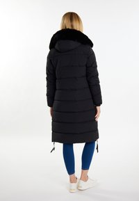 Sort pufferjakke med en pelsforet hætte, prydet med horisontale syninger og snørelukninger i kanten, parret med blå leggings og hvide sneakers.
