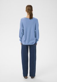 Világoskék pulóver puha anyagból, laza szabással és lehajtott vállakkal. Laza sötétkék farmernadrággal párosítva.