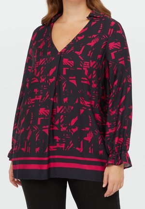 Femme portant un chemisier noir à manches longues avec un motif géométrique abstrait rouge et des poignets volantés, associé à un pantalon noir.