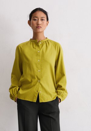 Limoengroene blouse met knopen en een Mandarin-kraag, met geplooide details op de schouders en elastische manchetten. Gepaard met zwarte broek.
