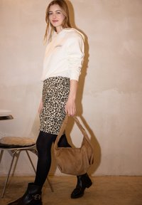 Femme portant un sweat à capuche blanc, une jupe imprimé léopard, des collants noirs et des bottes noires, tenant un sac à main en daim beige, marchant à l'intérieur.