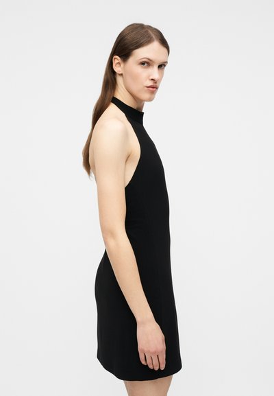 Νεαρή γυναίκα με μακριά καστανά μαλλιά, φορώντας μαύρο φόρεμα χωρίς μανίκια με λαιμόκοψη halter, στέκεται πλαγίως μπροστά σε απλό λευκό φόντο.