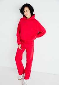 Hanorac roșu cu buzunar tip cangur și șnur, purtat cu pantaloni de trening roșii asortați. Material neted și croială lejeră, completat cu adidași argintii.