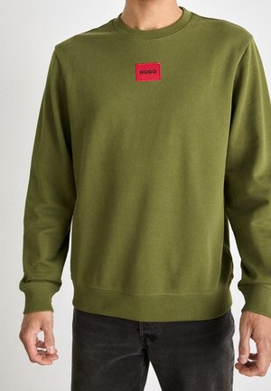 Homme portant un sweat à col rond vert olive avec un écusson logo rouge « HUGO » sur la poitrine, associé à un pantalon foncé.
