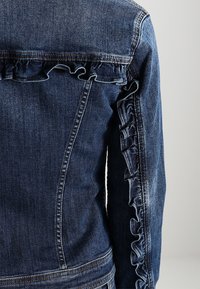 Jeanjacka i mörkblått med volangdetaljer längs axlarna och ärmarna. Har klassiska sömmar och en figursydd design.