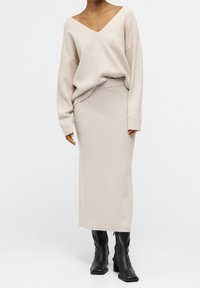 Pull beige en maille avec un col en V et des manches larges, associé à une jupe beige ajustée assortie. Bottes noires mi-mollet à talons blocs.