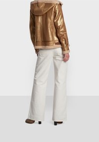 Gul metallisk jacka med huva, foder av fårskinn och manschetter, i kombination med vita byxor med vida ben och bruna klackskor. Bakvy.