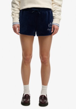 Person iført marineblå velourshorts med snøre, cremefarvet sweatshirt, hvide sokker og mørkebrune loafers står oprejst.