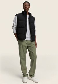 Juoda pūkinė liemenė su aukštu apykakliu, priekyje užtrauktuku ir tekstūruotomis skiltymis, dėvima ant šviesiai pilkos džemperio ir alyvuogių žalios spalvos kelnių. Balti sportbačiai.