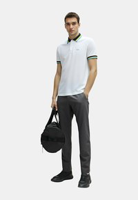 Polo bianco con colletto nero e dettagli a strisce verde-giallo-blu, abbinato a pantaloni grigio scuro e scarpe nere, mentre tiene una borsa rotonda.